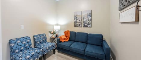 A room with a blue sofa, two patterned armchairs, a small table with a lamp, and wall art.