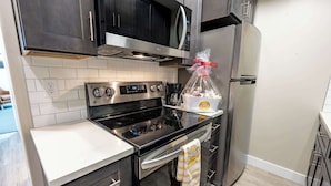 A modern kitchen with a stainless steel stove, oven, and microwave. There is a refrigerator and a bag of groceries on the counter.