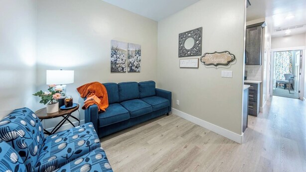 A living room with a blue sofa, a coffee table, and a lamp. There are two wall art pieces and a welcome sign. - Main St Suite | King Bed + Loft Bed | Full Kitchen (Pleasant Grove)