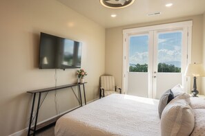 A bedroom with a bed, a TV mounted on the wall, a chair, a bedside table with a lamp, and a window with a view. - Upscale 3BR Retreat-Spacious Living - Waffles too! (Pleasant Grove)