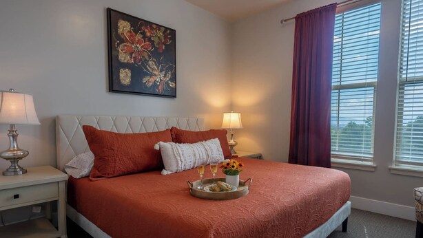 A hotel room with a large bed, two bedside lamps, a painting on the wall, and a window with curtains.