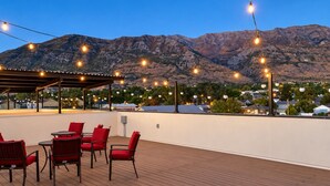 A rooftop patio with a wooden deck, red chairs, and string lights against a mountainous backdrop.