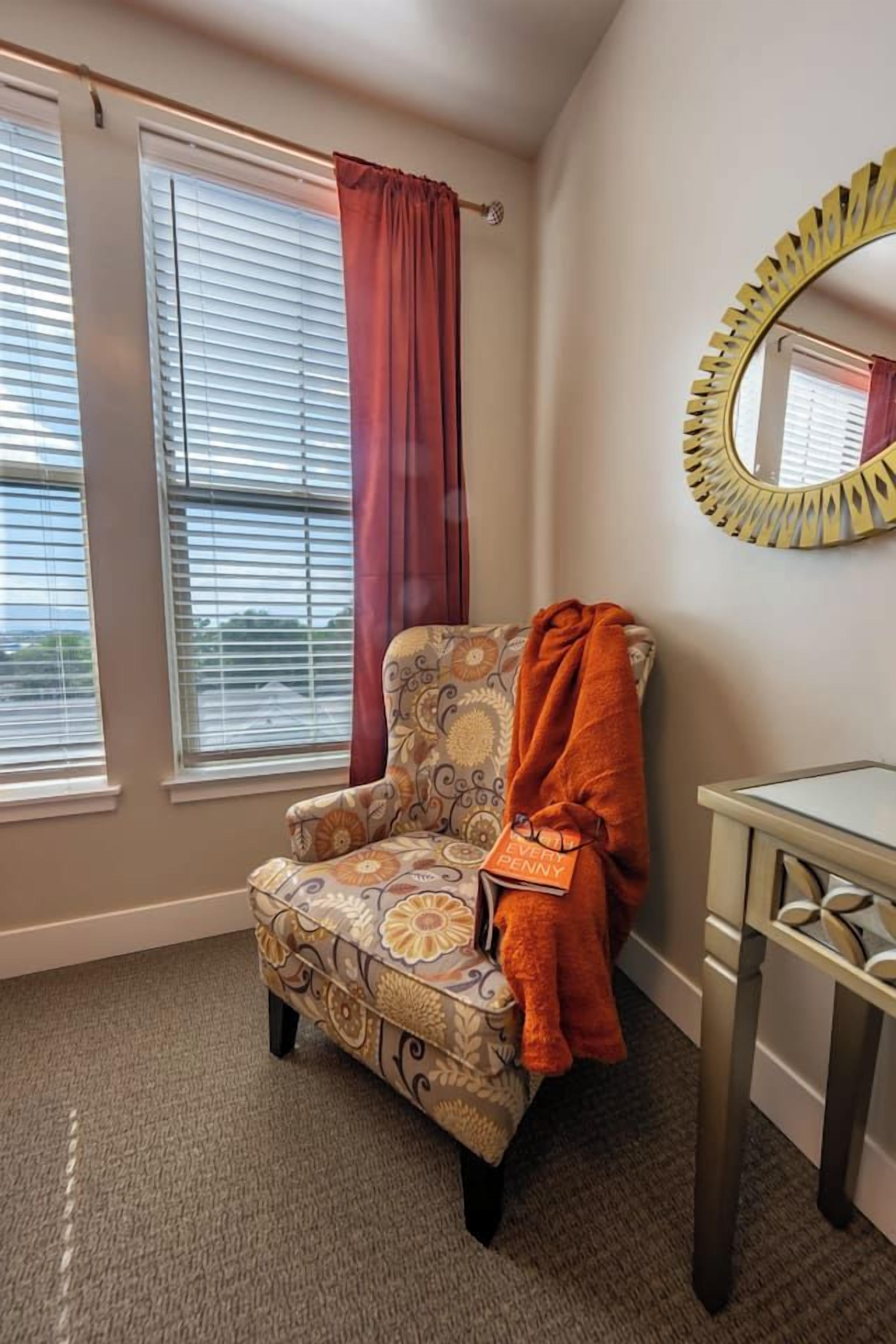 A room with a patterned armchair, a mirror with a yellow frame, and a window with blinds.