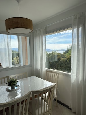Een eetgedeelte met een witte tafel en stoelen, een hanglamp en een uitzicht op een natuurlandschap door grote ramen.