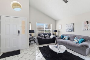 A living room with a black sofa, a grey sofa, a white coffee table, a black rug, a white door, and a window with blinds. - Spacious 4-bedroom Pool house in charming Kissimmee Close to Disney  with Wi-fi (Kissimmee)