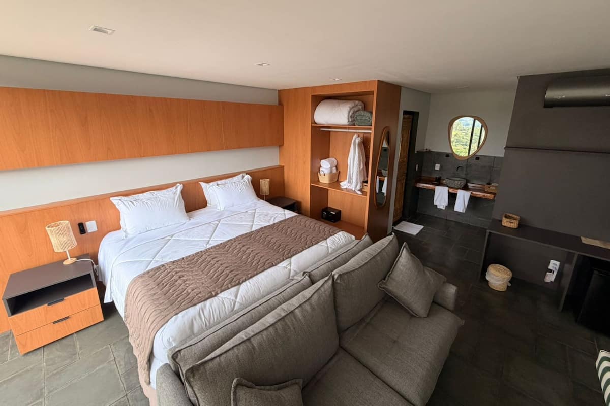 Una habitación de hotel moderna con una cama grande, cabecera de madera, armario empotrado, mesita de noche pequeña, espejo redondo, sofá de tonos oscuros y un baño con pared de piedra y lavabo.