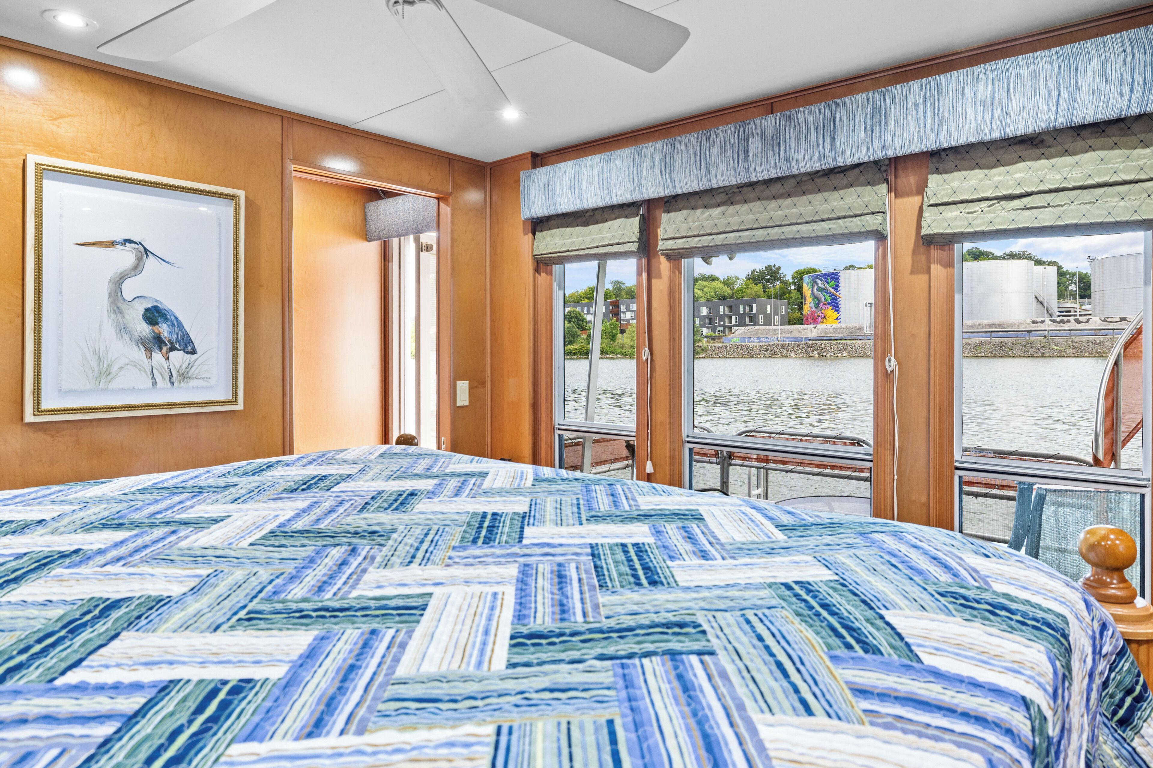 A room with a large bed, a framed picture of a bird, and a view of a body of water through large windows.