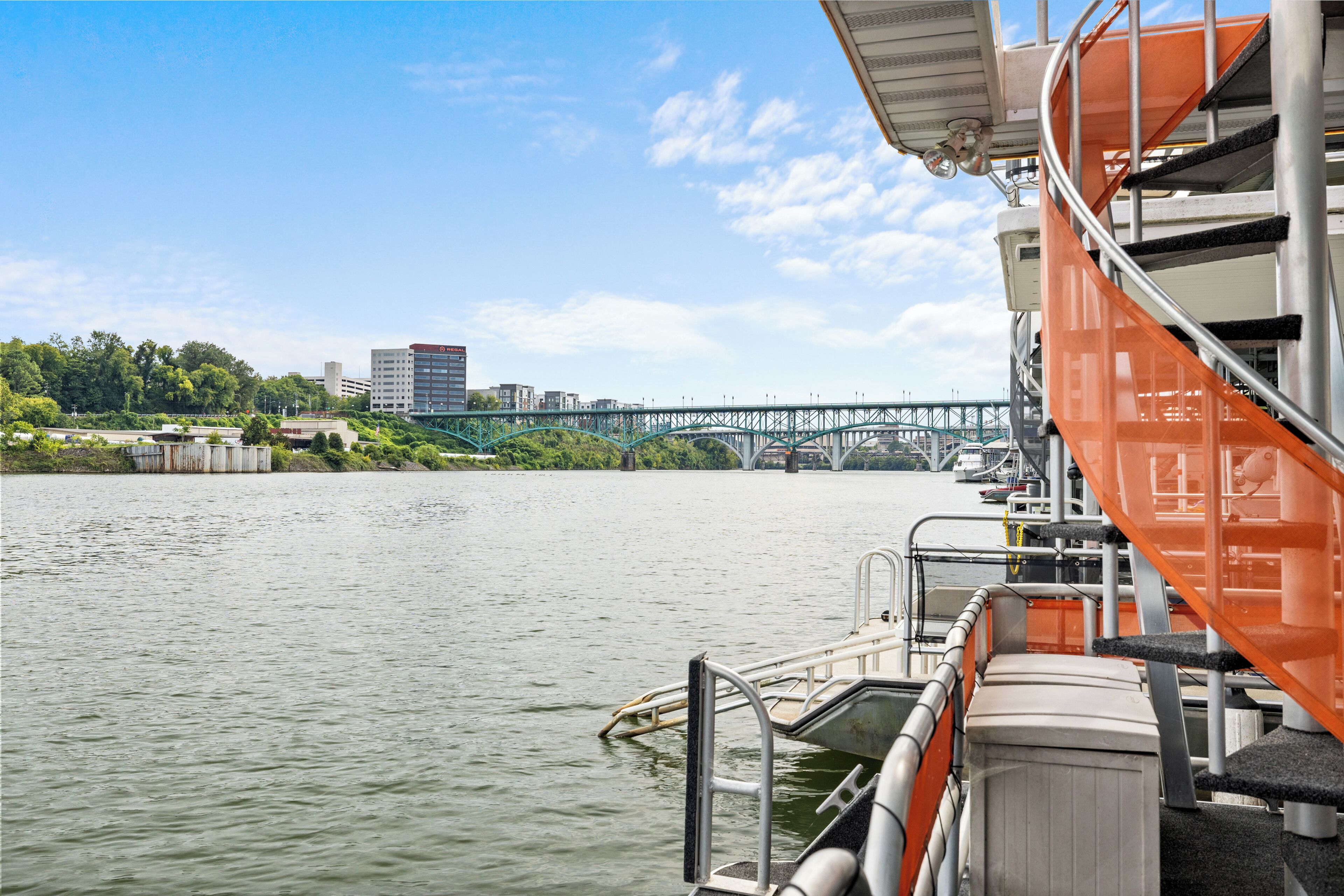 Un río con un puente al fondo, una barca amarrada con barandillas naranjas, y un edificio de techo inclinado.