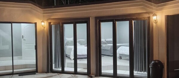 Una habitación de hotel con un amplio ventanal corredero, una cama con cabecera gris y una mesita con un jarrón.