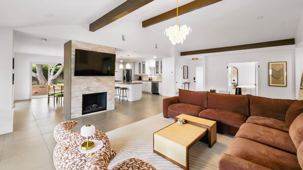 Un salón amplio con un sofá marrón grande, una mesa de centro y un reposapiés estampado. Se observa una chimenea al fondo, y la habitación tiene un diseño moderno con vigas a la vista y un televisor grande montado en la pared.