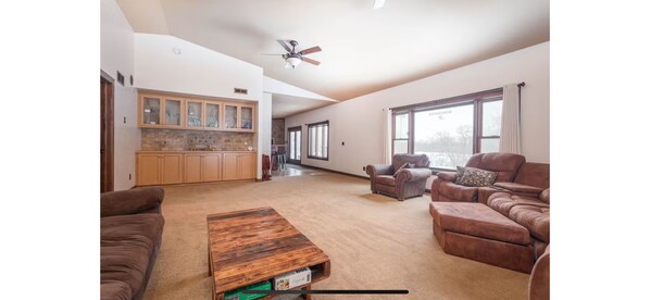 A spacious living room with a brown leather sofa, a wooden coffee table, and a view of the kitchen and dining area through large windows. - Private 40-Acre Cabin + Indoor Pickleball & Trails (Sauk Centre)