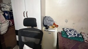 Une petite pièce avec une armoire blanche, un fauteuil de bureau noir, une petite table avec un radiateur et un lit avec une literie à motifs.