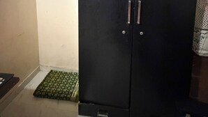 Une pièce avec une armoire de couleur sombre, un petit tapis vert sur le sol et un rideau blanc orné d’un motif floral.