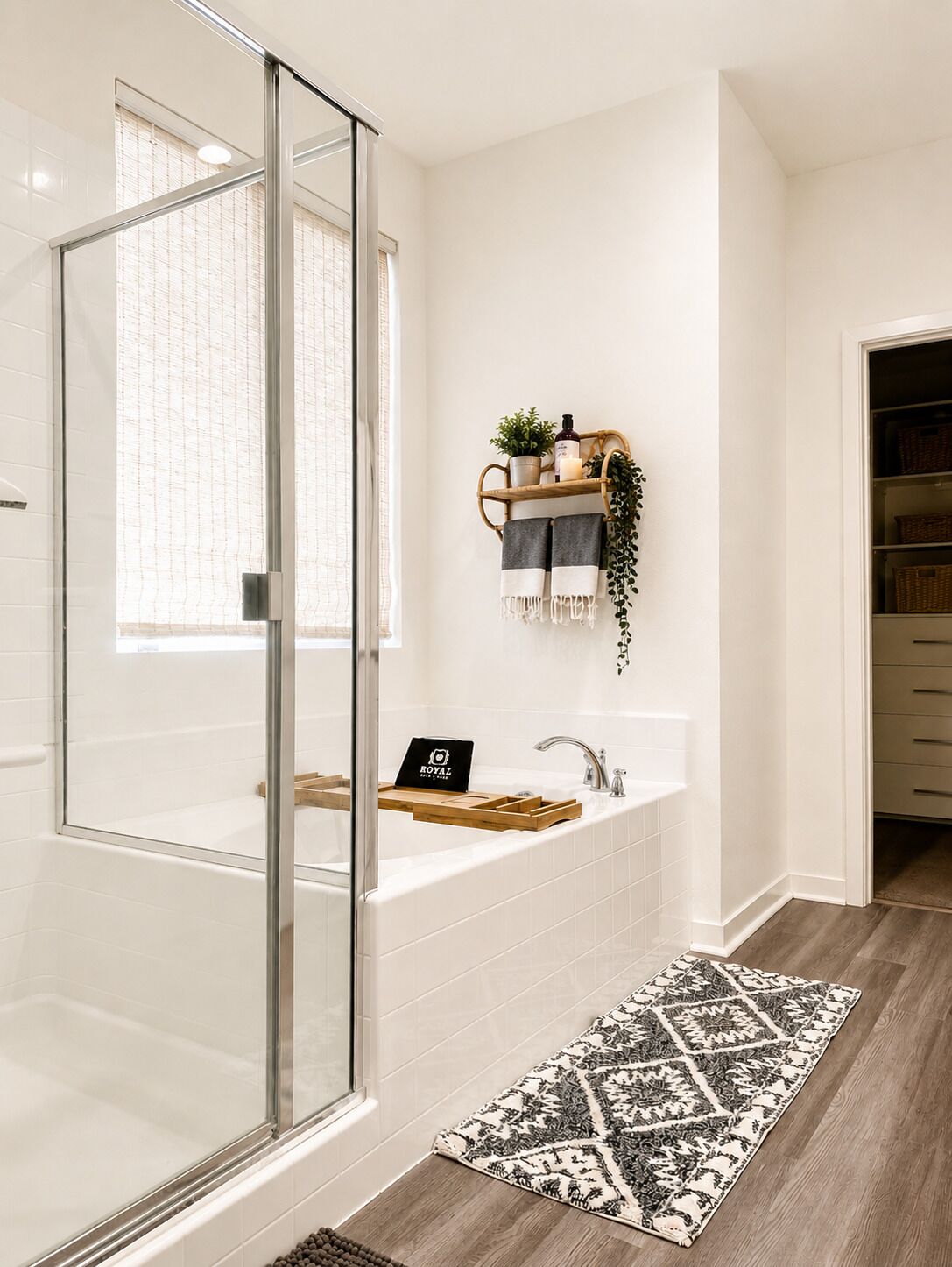 A bathroom with a glass shower enclosure, a white bathtub, a wooden shelf with toiletries, and a patterned rug on the floor.