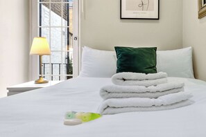 A neatly made bed with white linens, a green pillow, and a stack of folded towels. - Stylish 2BR Kensington Retreat, 1 min to Station! (Greater London)