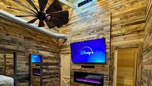 Una habitación al estilo de cabaña de troncos con una cama, ventilador de techo, televisión montada en la pared y chimenea.
