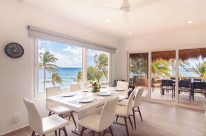 A dining area with a view of the ocean, a clock on the wall, and a large sliding glass door leading to an outdoor seating area. - Casa Lindavista | 3BR | Condo | Puerto Aventuras (Puerto Aventuras)