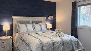 Ein ordentlich bezogenes Bett mit gestreifter Bettdecke, Nachttischlampen, einem Nachttisch mit Lampe und einem Fenster mit Vorhängen.