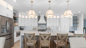 Uma cozinha moderna com armários brancos, uma ilha central com banquetas, eletrodomésticos de aço inoxidável e iluminação pendente.