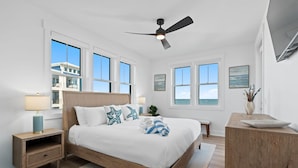 Quarto com cama de madeira, paredes brancas, janelas amplas, ventilador de teto e vista para o oceano.