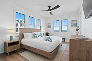 A bedroom with a wooden bed, white walls, large windows, a ceiling fan, and a view of the ocean.