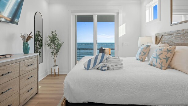 Quarto com cama, penteadeira, televisão, planta e vista para o mar.