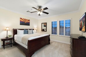 Ein Schlafzimmer mit einem Bett, einem Deckenventilator, einem Nachttisch mit Lampe, einer Kommode, einem Fenster mit Jalousien und einem Bild an der Wand.