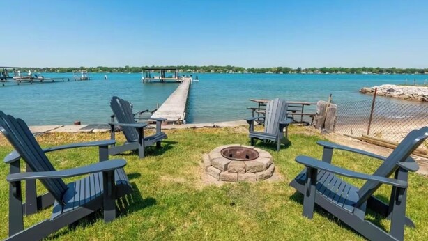 Una terraza junto al lago con sillas Adirondack, una fogata y un muelle que se adentra en el agua.