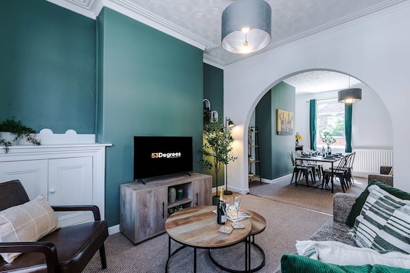A living room with teal walls, a wooden coffee table, a TV, and a dining area with a table and chairs. - Stylish 2-bed House in Crewe Perfect for Business (Crewe)