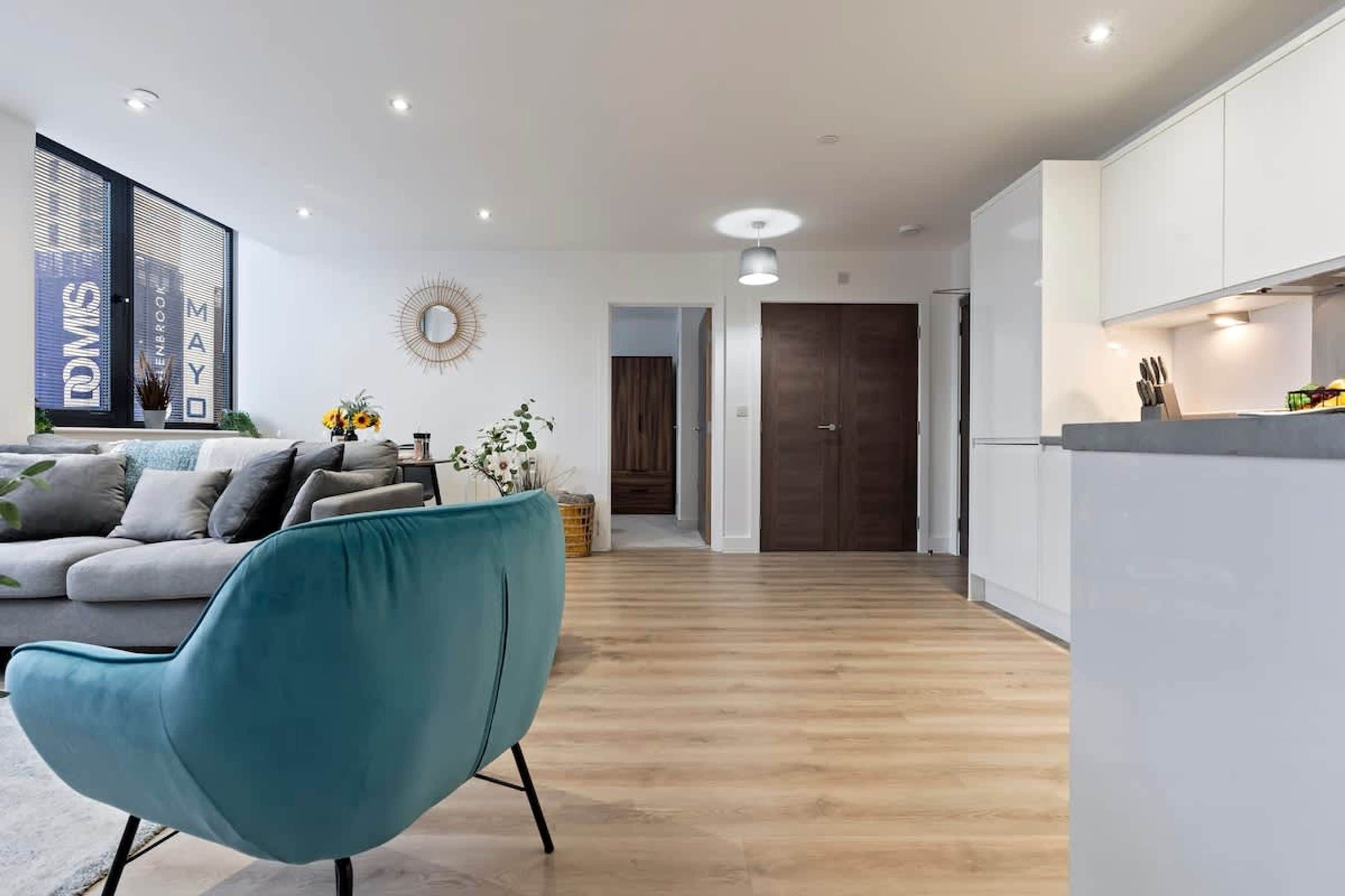 A modern living room with a sofa, teal chair, and a kitchen area with cabinets and a countertop.