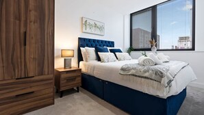 A bedroom with a large wardrobe, a bed with blue bedding, a bedside table with a lamp, and a window with blinds. - Pristine Flat Ideal for Small Groups Sleeps 4 (Manchester)