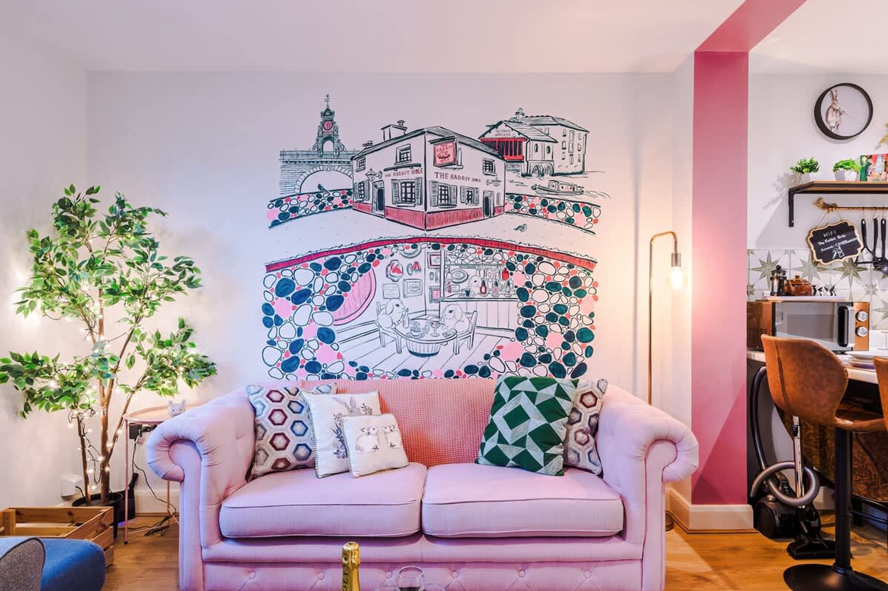 Ett mysigt vardagsrum med en rosa soffa, en väggmålning, en växt och en köksdel med en mikrovågsugn.