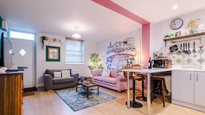 A living room with a grey sofa, a pink sofa, a coffee table, and a kitchen area with a white cabinet and a hanging pot rack. - Stunning City Centre Home Perfect For Couples (Chester)