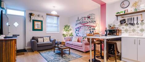 A living room with a grey sofa, a pink sofa, a coffee table, and a kitchen area with a white cabinet and a hanging pot rack.