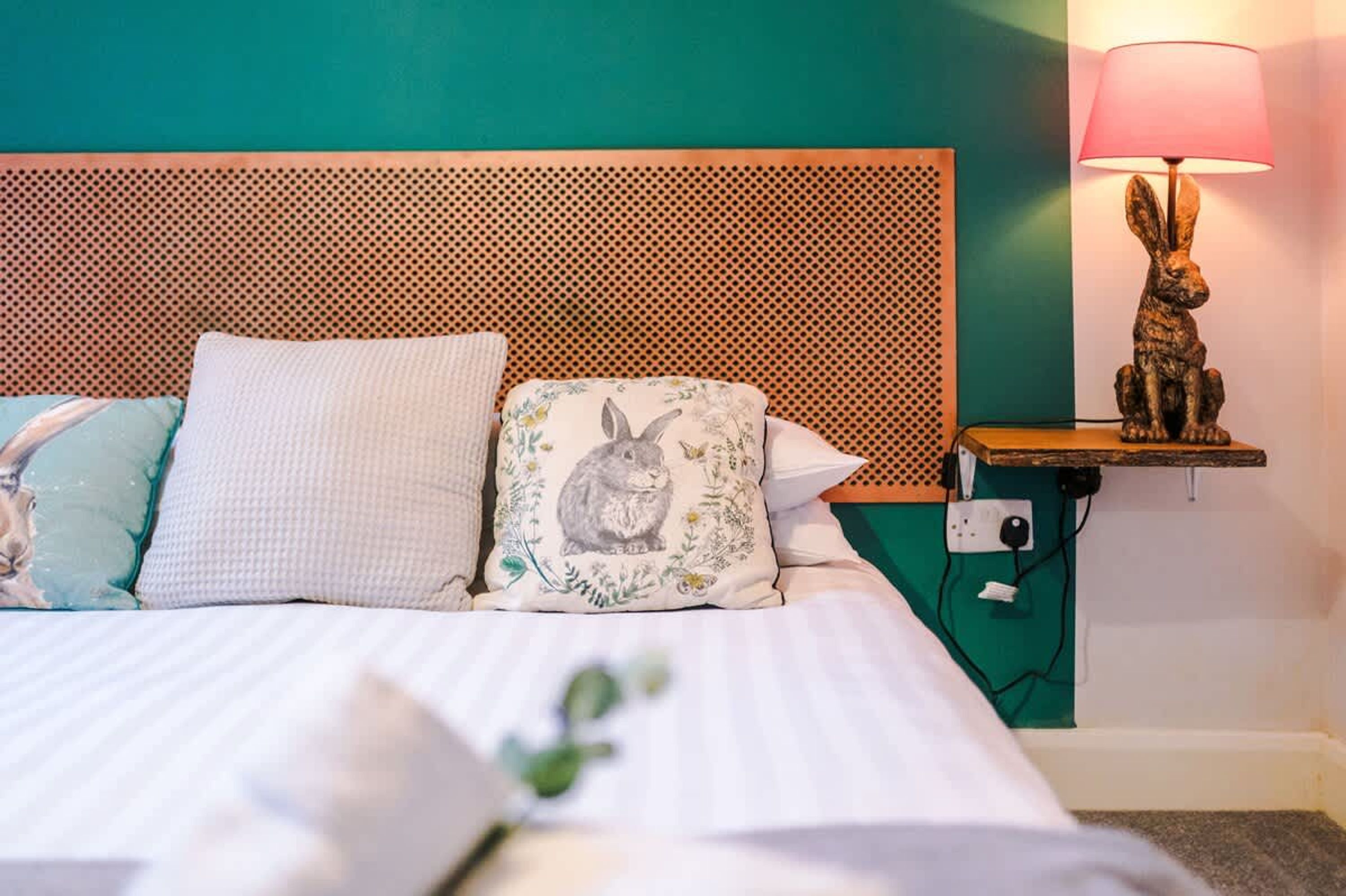 A bed with patterned pillows, a wooden headboard, a wall-mounted lamp, and a power outlet.