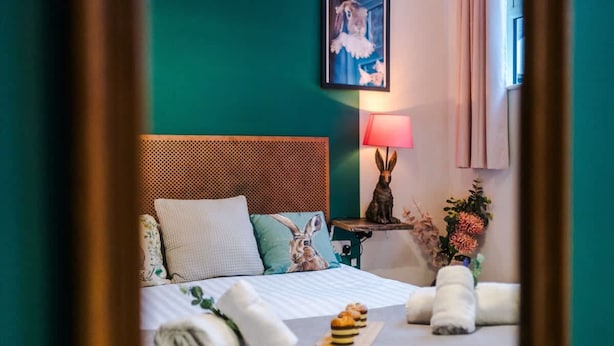A neatly made bed with pillows, a bedside lamp, a framed picture, and a small table with a vase of flowers. - Stunning City Centre Home Perfect For Couples (Chester)