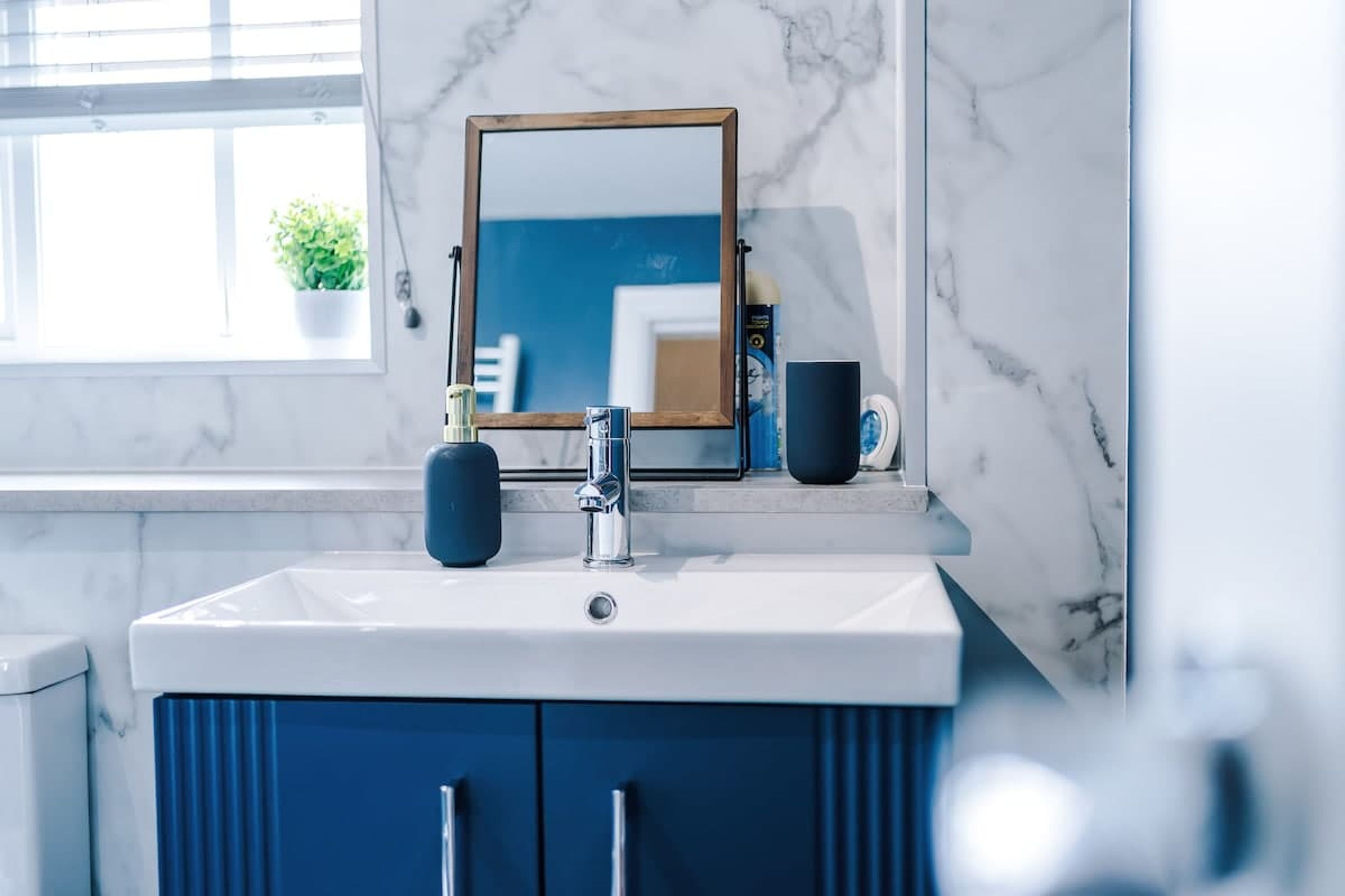 A bathroom with a white sink, dark blue cabinets, a wooden-framed mirror, and a window with a potted plant.
