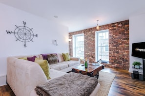 A modern living room with a brick wall, a white sofa, a coffee table, and a television. - NEW Stunning City Centre Flat Amazing Location (Chester)
