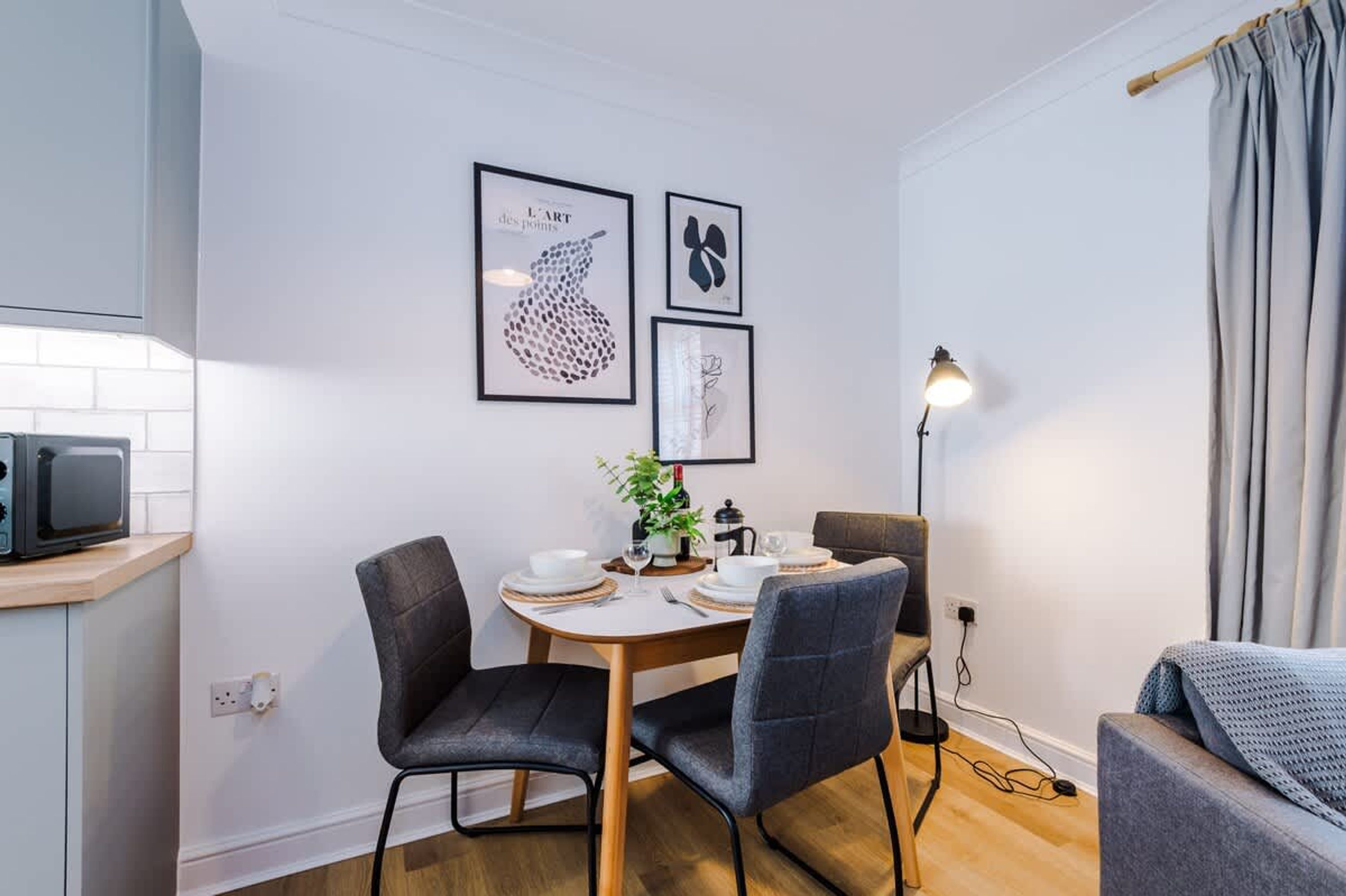 A modern dining area with a round table, two chairs, a small plant, and framed artwork on the wall.