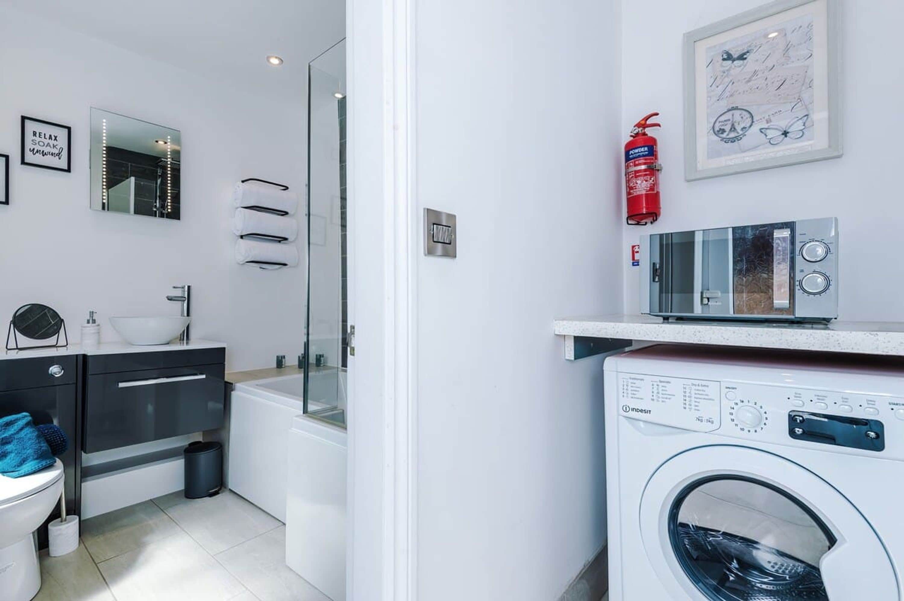 A modern bathroom with a glass shower enclosure, a white sink, a black vanity, a white toilet, and a wall-mounted washing machine.
