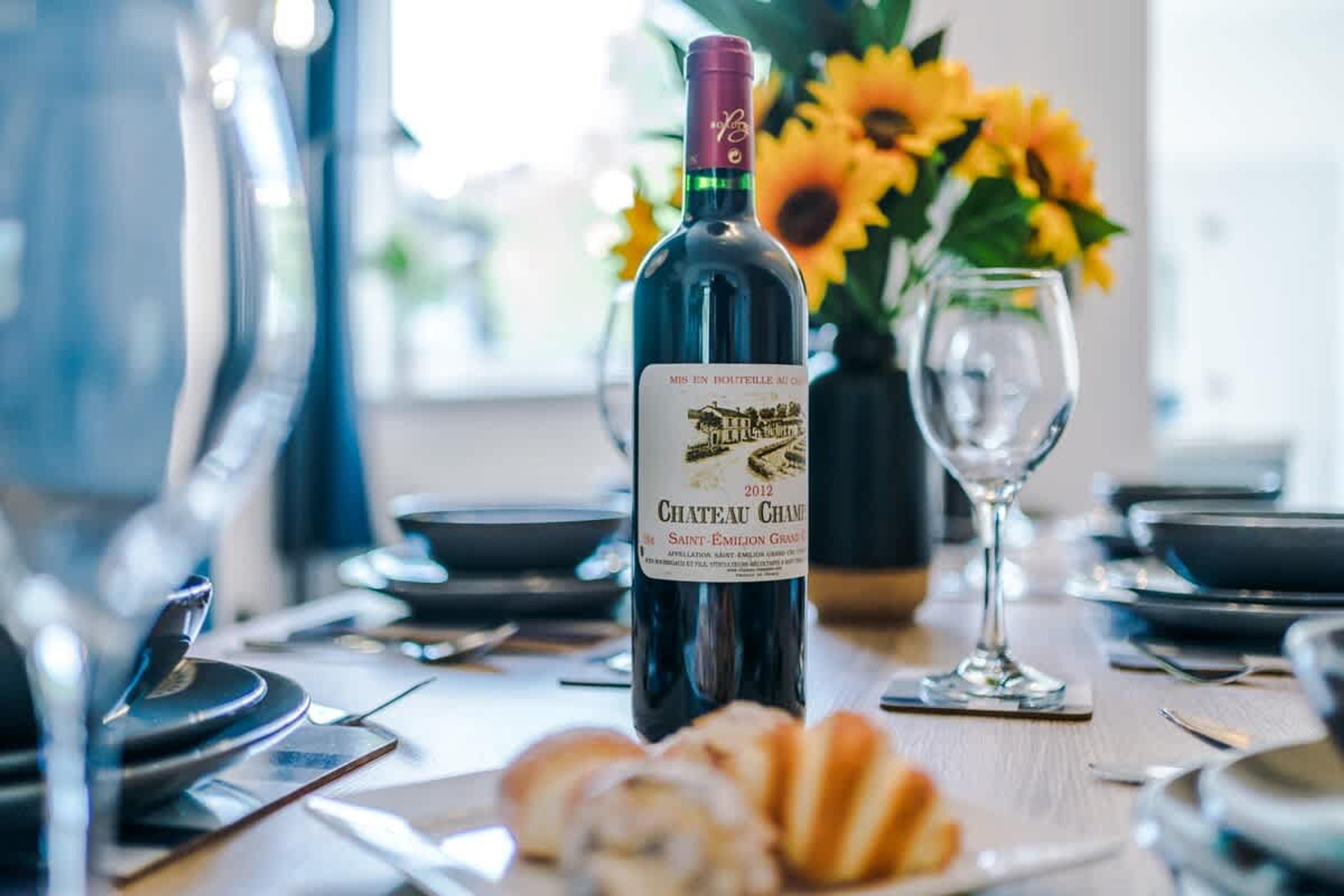 A dining table with a bottle of Chateau Chanteur wine, a glass, and a vase with sunflowers.