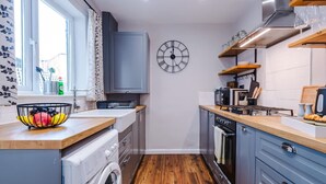A compact kitchen with a washing machine, a clock, and a window with curtains. - NEW Spacious Stylish Home City Centre Sleep 4 (Chester)