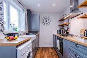 A compact kitchen with a washing machine, a clock, and a window with curtains. - NEW Spacious Stylish Home City Centre Sleep 4 (Chester)