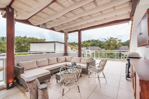 Uma área de estar coberta ao ar livre, com sofá, cadeiras de vime e uma mesa.
