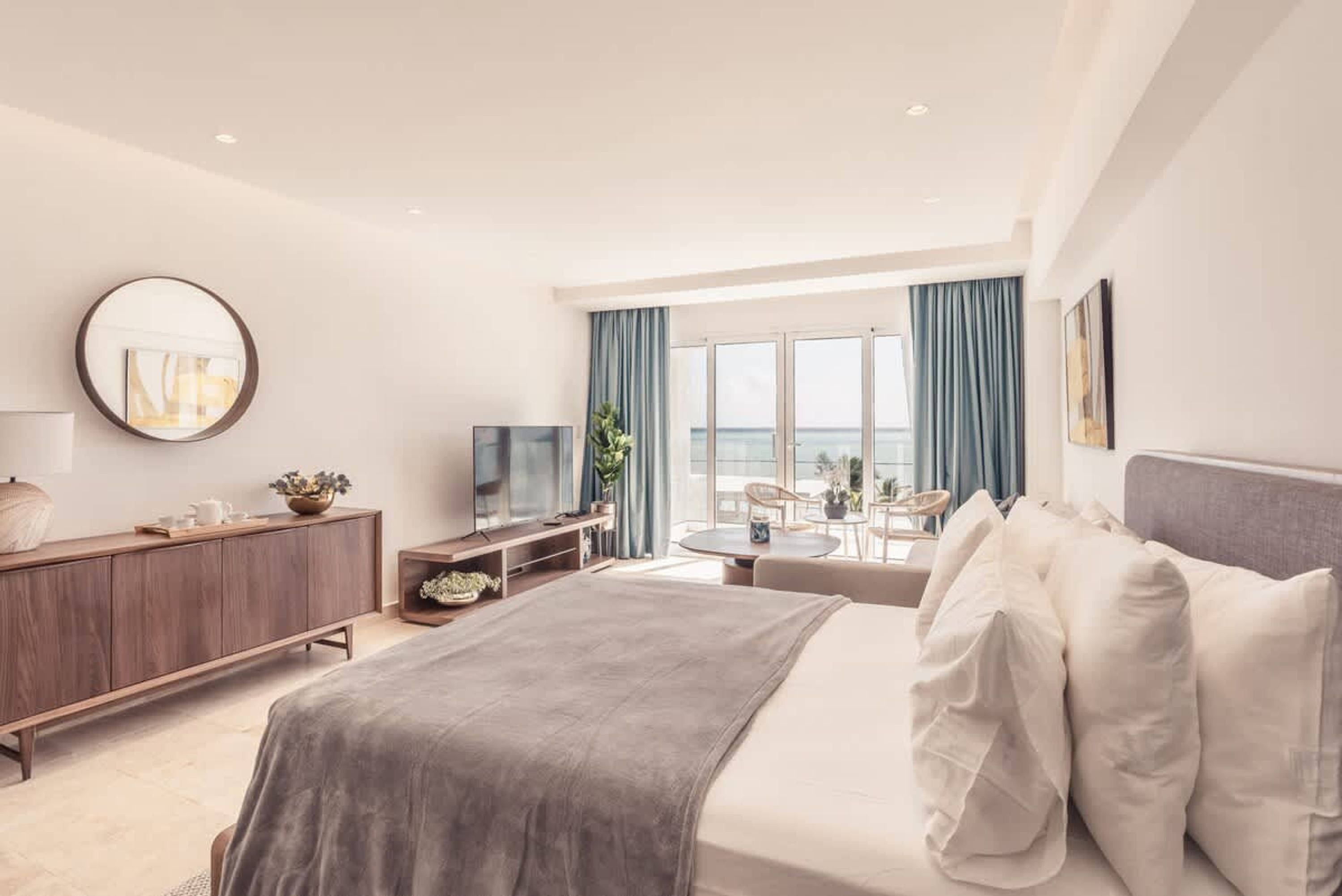 A modern hotel room with a bed, a TV, a desk, and a view of the sea.