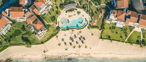 An aerial view of a resort with a central swimming pool, beachfront, and multiple buildings with red-tiled roofs.