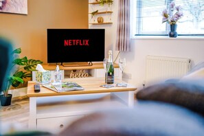 A living room with a TV displaying 'NETFLIX,' a wooden coffee table with a bottle, glasses, and a remote control, a 'hello' sign, and a potted plant. - Spacious House Incredible Location Free Parking (Chester)