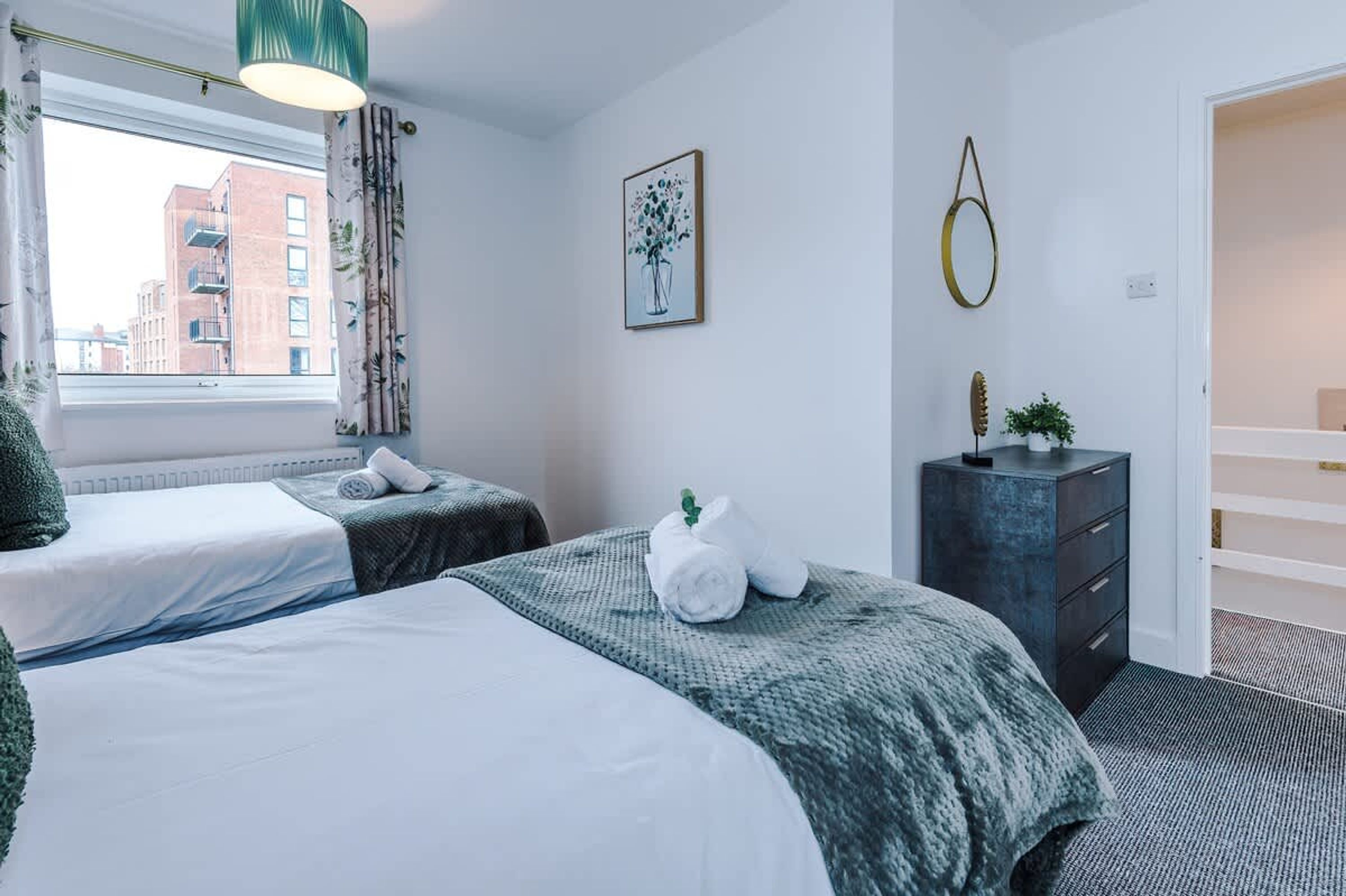 A hotel room with two beds, a dresser, a framed artwork, and a view of buildings outside the window.