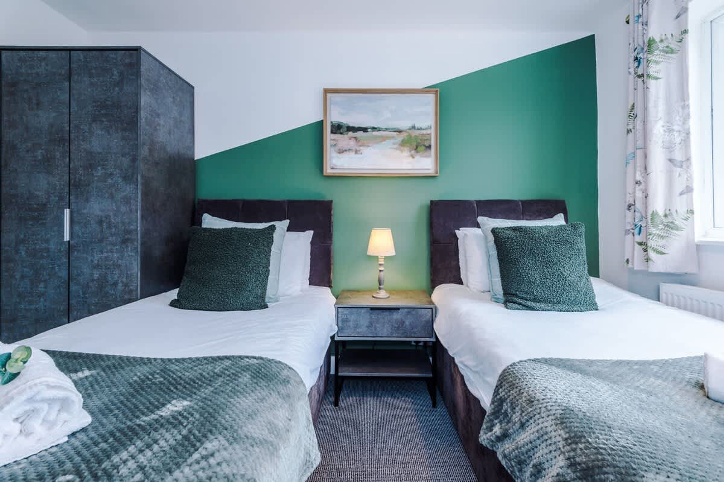 A hotel room with two beds, a nightstand with a lamp, a painting on the wall, and a wardrobe.
