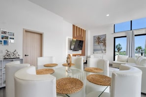 Un salon moderne avec une table basse en verre, des canapés blancs et un téléviseur fixé sur un panneau en bois.